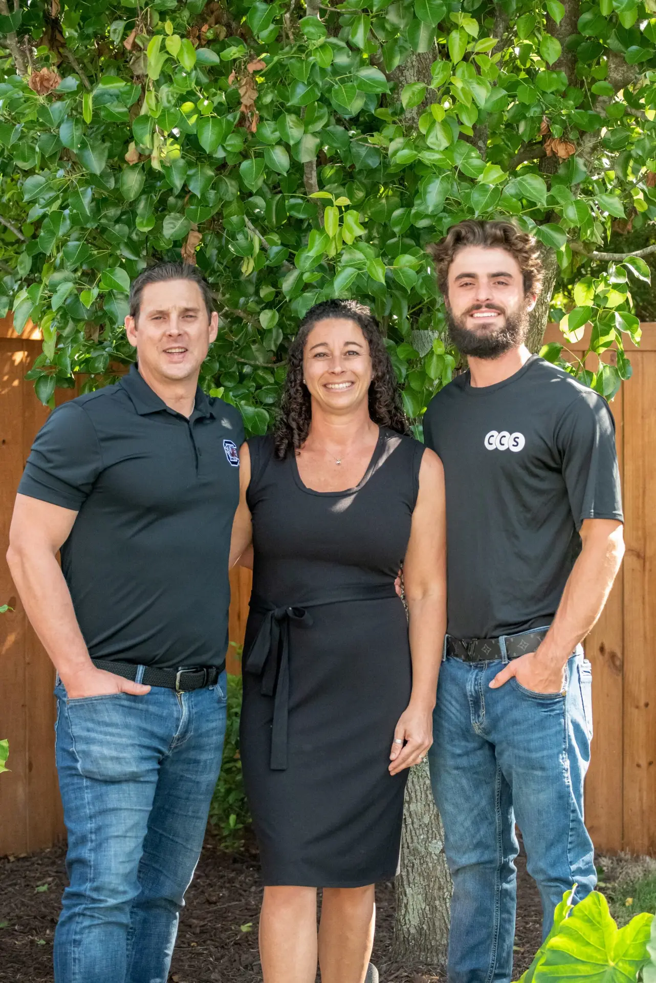 Three professionals in black shirts posing together against a green ivy backdrop with wooden fence