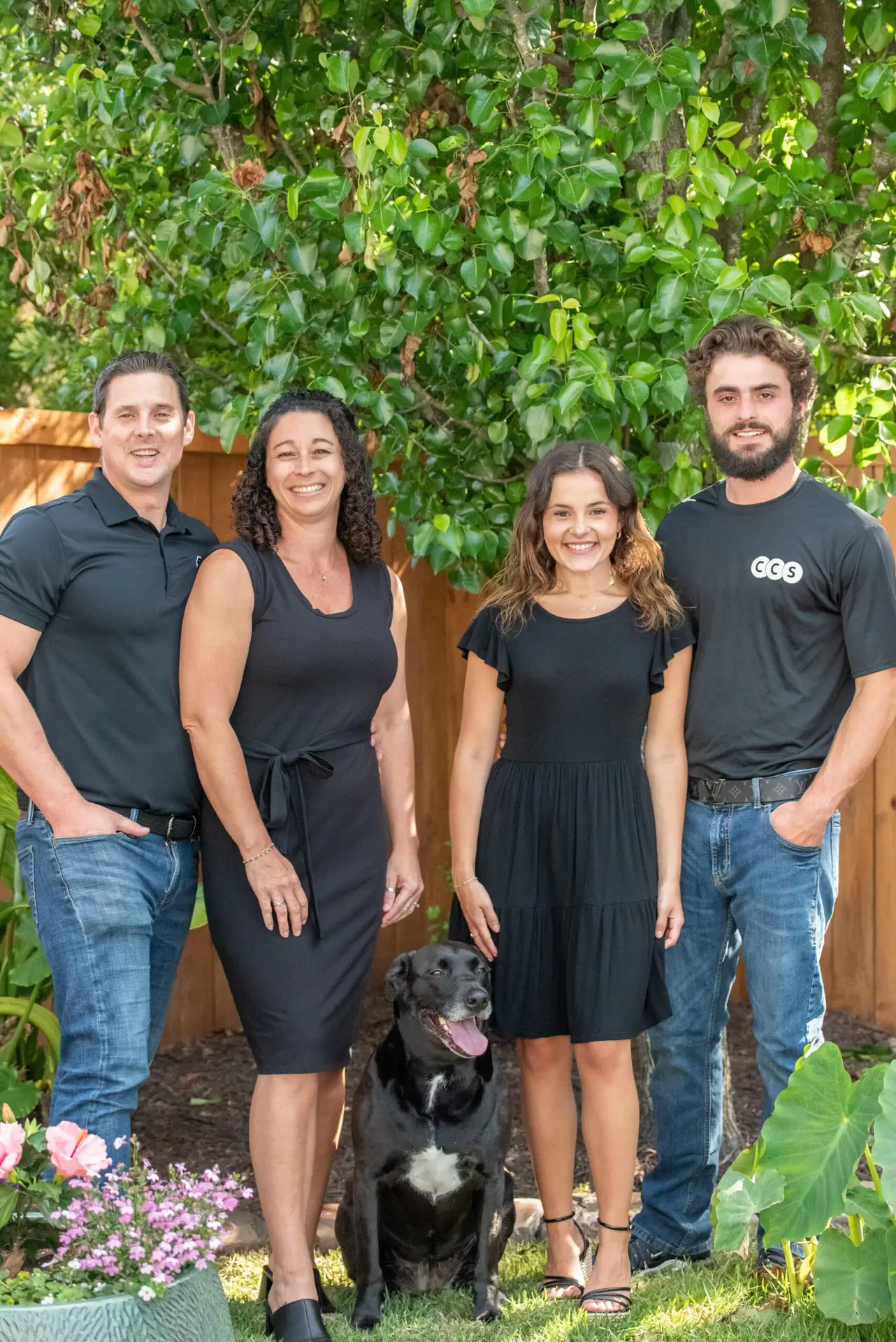 Four people in black GGG branded shirts posing with a black dog in a garden setting