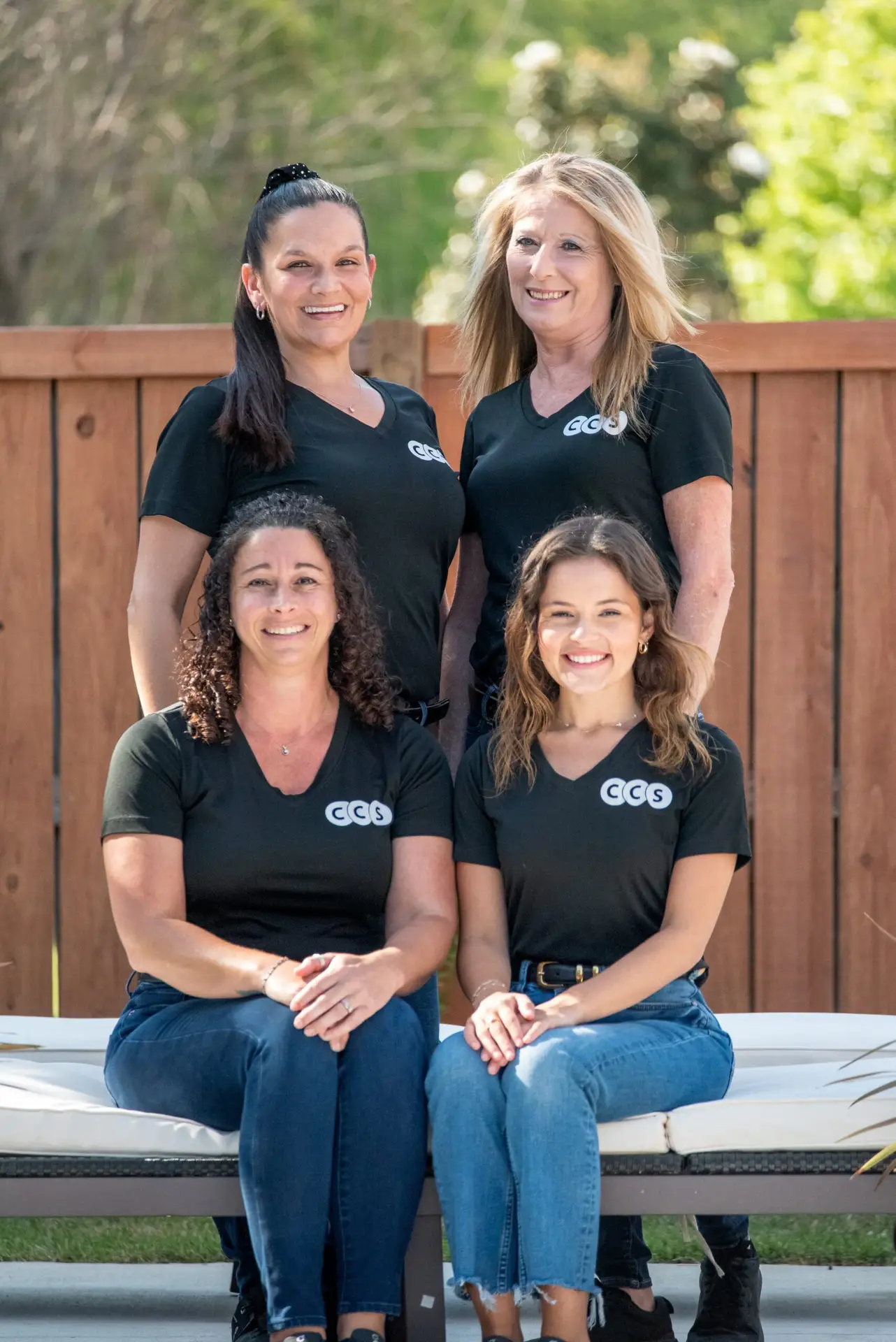 Four women wearing black GGG branded shirts seated and standing against a wooden fence backdrop